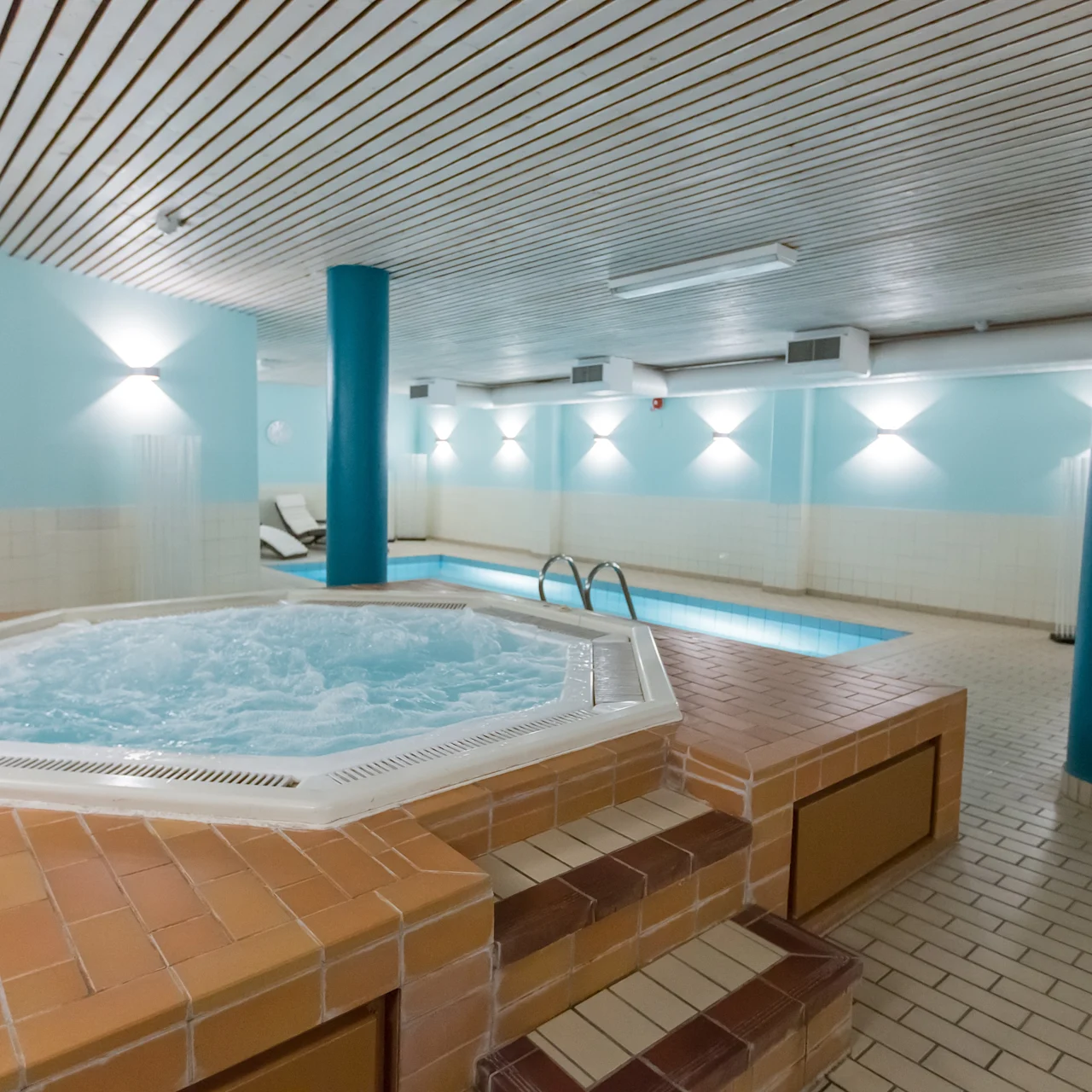 A hot tub bubbles in the foreground, surrounded by brick tiles. In the background, an indoor swimming pool is seen in a softly lit room with light blue walls.