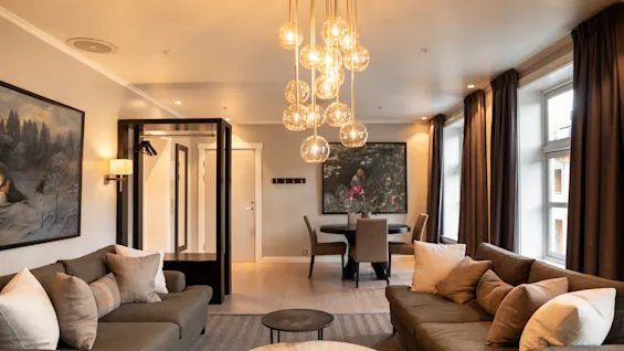 Luxurious living room features a contemporary chandelier illuminating two sofas with cushions, centered by a round coffee table. Art adorns walls; windows draped with curtains provide natural light.