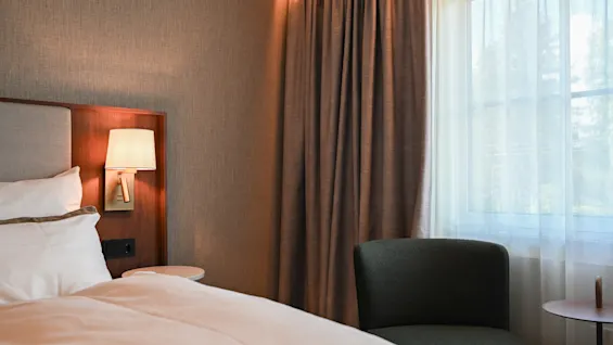Et koselig hotellrom med en komfortabel seng, en varm nattbordslampe og en stol ved siden av et vindu med gardiner.