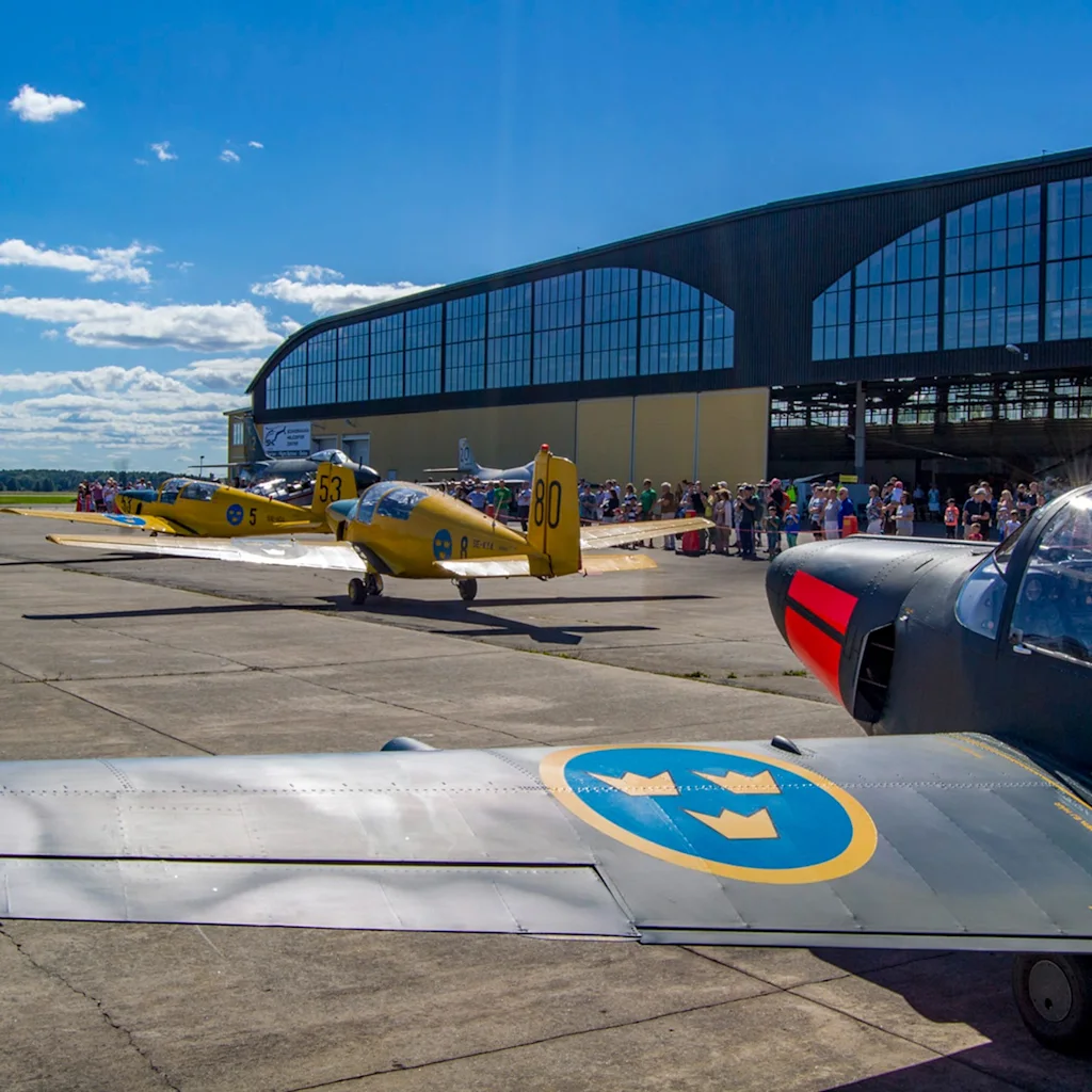 Airplanes on display at Västerås flygmuseum with a crowd of people and a large hangar under a blue sky.
