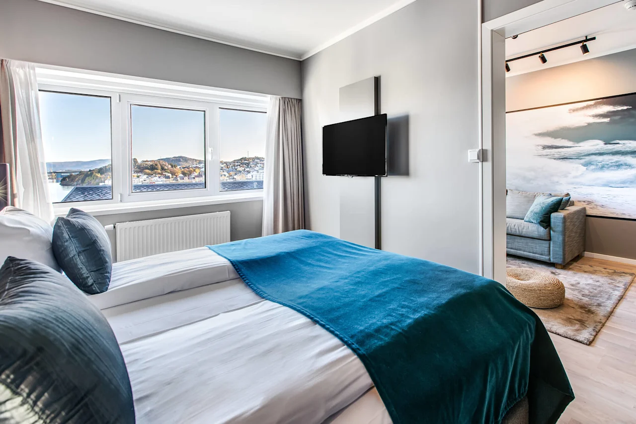 A neatly made bed with teal bedding faces a wall-mounted TV in a modern bedroom. Large windows display a scenic view. Adjacent room has sofa and ocean-themed decor.