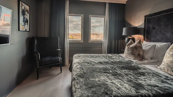 A bed with plush pillows sits in a cozy, dark-colored room. Large windows show a cityscape outside, and a chair and lamp complete the setting.