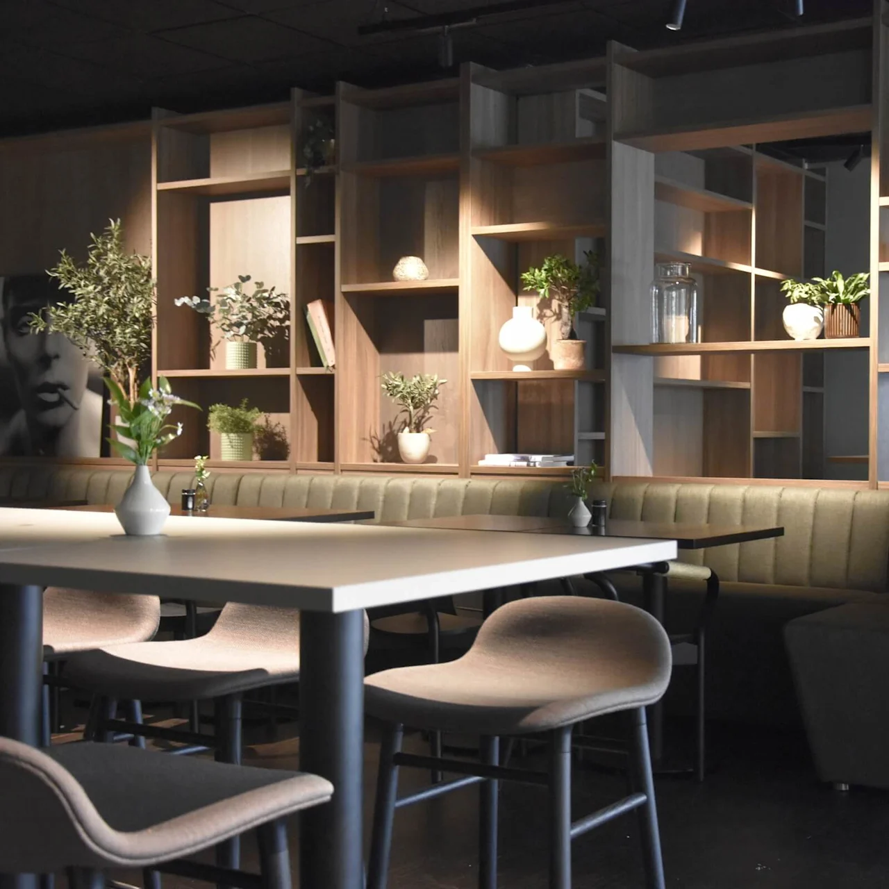 Modern dining area features a white table and chairs, surrounded by wooden shelving filled with plants and decor, against a dark background with a large green sofa and artwork.