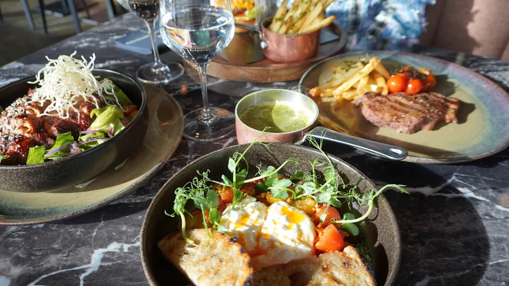 Ett bord med tre tallrikar mat, inklusive en sallad med krispig garnering, en stek med pommes frites och en skål med rostat bröd och grönsaker. Glas med vatten och vin följer med.