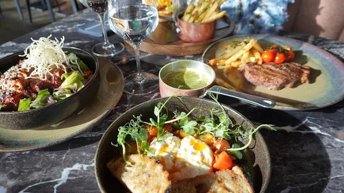 Pöytä, jossa on kolme lautasta ruokaa, mukaan lukien salaatti rapealla koristeella, pihvi ranskalaisilla ja annos paahtoleivällä ja kasviksilla. Mukana vesilasit ja viini.