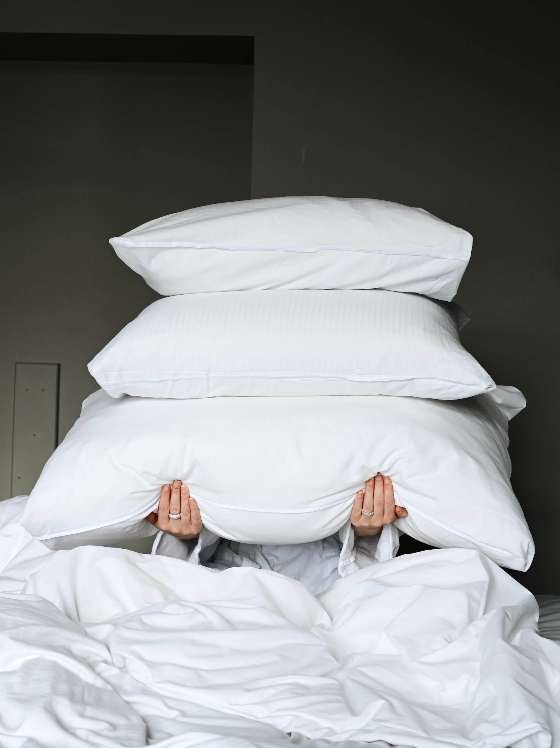 Person covered by a stack of pillows, holding the bottom pillow, in a bed with white linens against a grey wall.