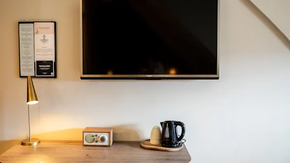 En väggmonterad TV är avstängd ovanför ett skrivbord av trä med lampa, retro radio, vattenkokare och koppar i en prydlig och modern hotellrumsmiljö.