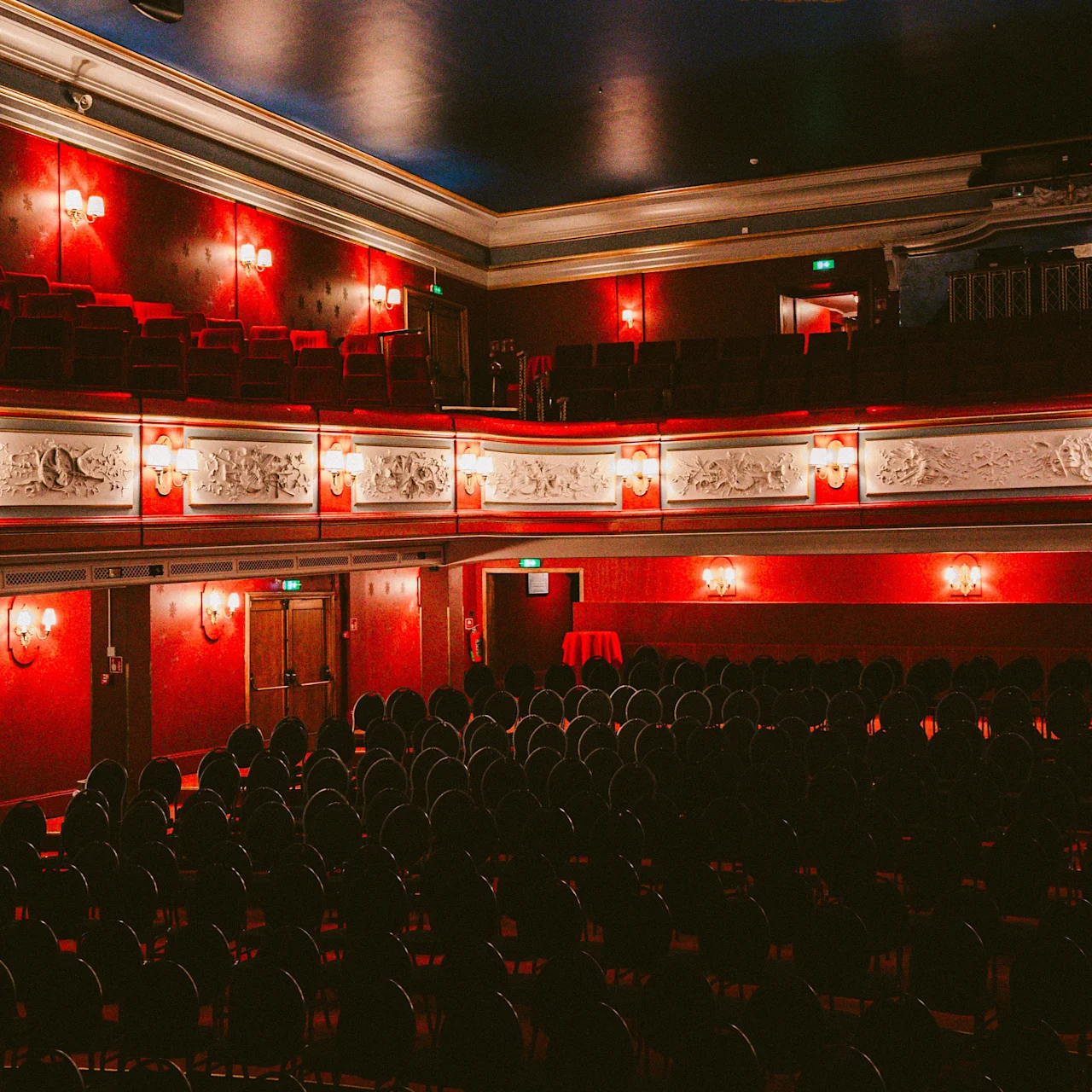 Rows of empty chairs face a stage in an ornate theater. A large chandelier hangs from the ceiling, with red and gold decor enhancing the elegant interior.