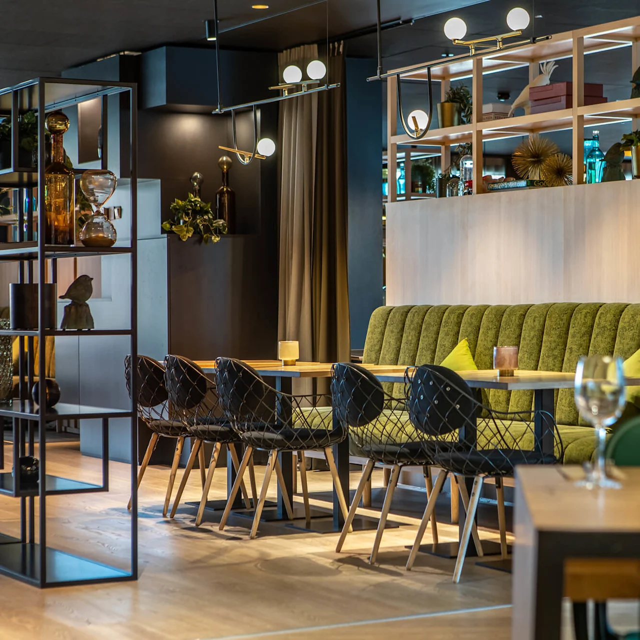 Dining tables adorned with elegant chairs surround a booth with green upholstery; modern lighting fixtures hang above, and decorative shelves display plants and glassware. Sophisticated restaurant ambiance.