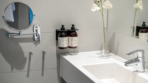 A mirror is mounted on a bathroom wall with two bottles of soap and a tall vase with white flowers beside a sink. The wall tiles are gray.