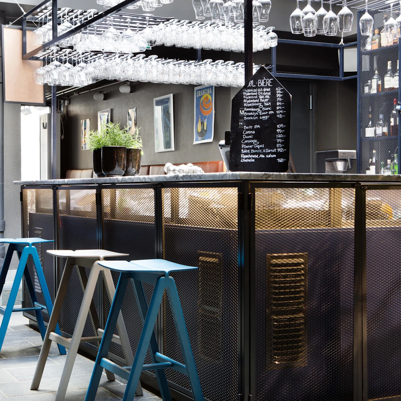 En stilfull bar har hengende glass, fargerike stoler og en tavle med forskjellige øl. Planter og kunst dekorerer det moderne rommet.