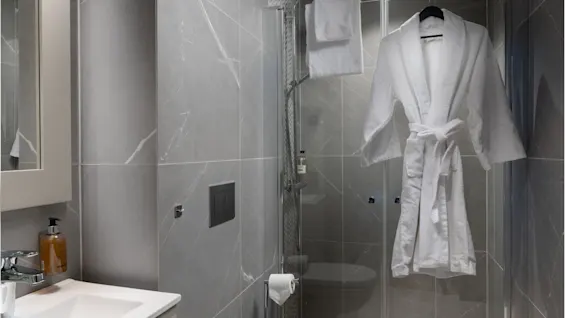 En hvid badekåbe hænger på en brusedør i et moderne badeværelse med grå marmorklinker, en vægmonteret toilet og en vask.