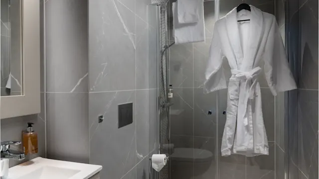 A white bathrobe hangs on a shower door in a modern bathroom with gray marble tiles, a wall-mounted toilet, and a vanity sink.