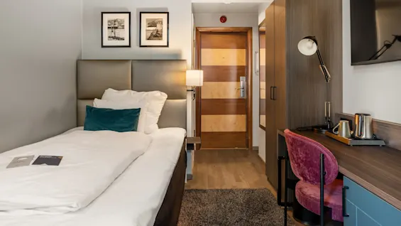 En seng med hvidt sengetøj og en turkis pude står nær en trædør på et hotelværelse. Vægkunst, et klædeskab og et skrivebord med en lampe skaber en hyggelig atmosfære.