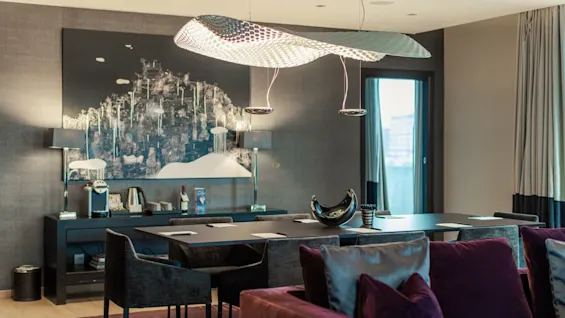 A modern dining room features a large, abstract ceiling light illuminating a dark table with chairs. The room includes decorative art, and a window with curtains offers natural light.
