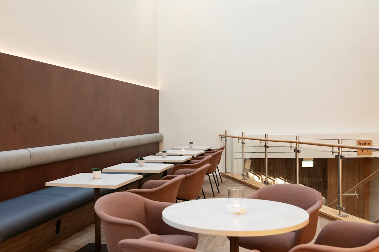 A modern cafe or lounge with comfortable seating, tables, and a railing overlooking a lower level. A sign reads Toaletter.