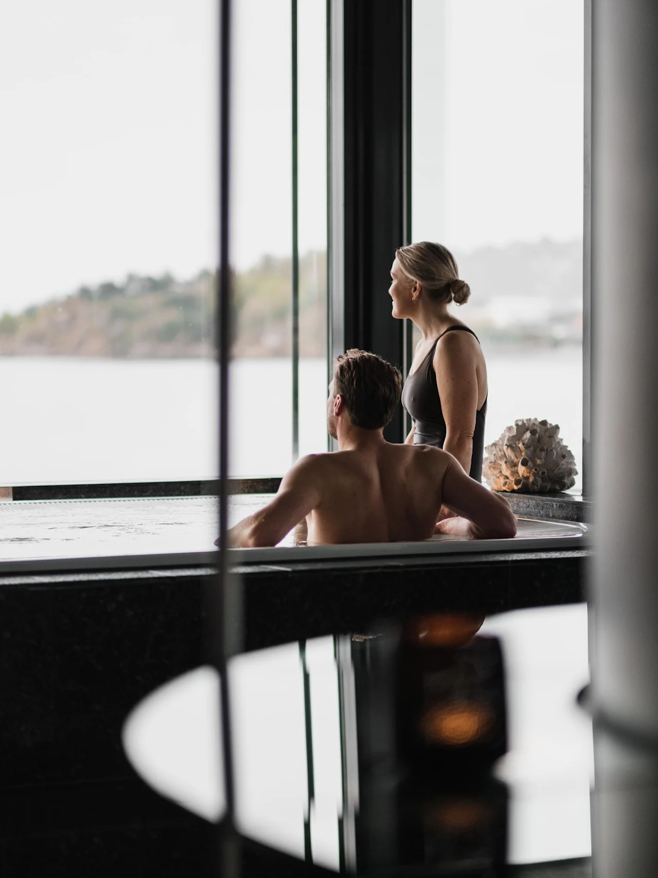 To personer slapper av i et boblebad, ser ut et stort vindu mot en naturskjønn utsikt over vann og fjerntliggende land.