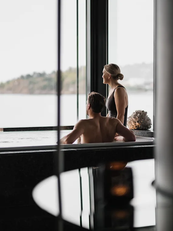 Två personer kopplar av i en bubbelpool och tittar ut genom ett stort fönster mot en naturskön utsikt över vatten och avlägsen mark.