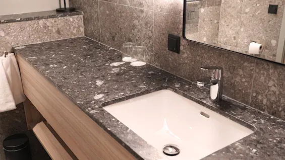 A sink with a metallic faucet is set into a sleek, dark marble countertop. Towels and glass cups are nearby, against a stone-tiled backsplash in a modern bathroom.