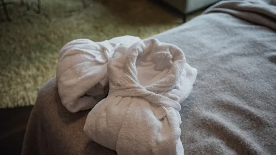 Kaksi pehmeää valkoista kylpytakkia siististi taiteltuna sängyllä, kutsuen mukavuuteen ja rentoutumiseen viihtyisässä ympäristössä.