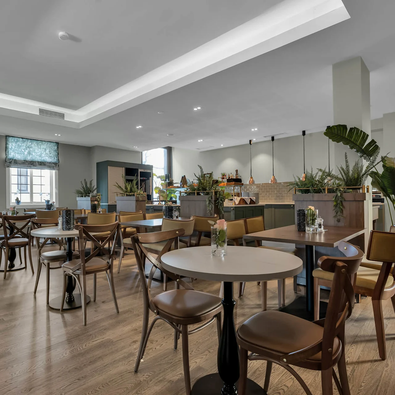 Ett modernt caféinteriör med bord, stolar och en bardel; stora fönster och inomhusväxter förstärker den luftiga atmosfären.
