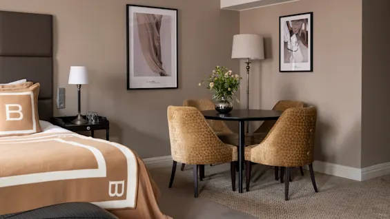 Seng med brunt sengeteppe og "B" initial ved siden av en lampebelyst nattbord; nært rundt spisebord med vase med blomster og fire stoler; innrammet billedkunst pryder beige rom.