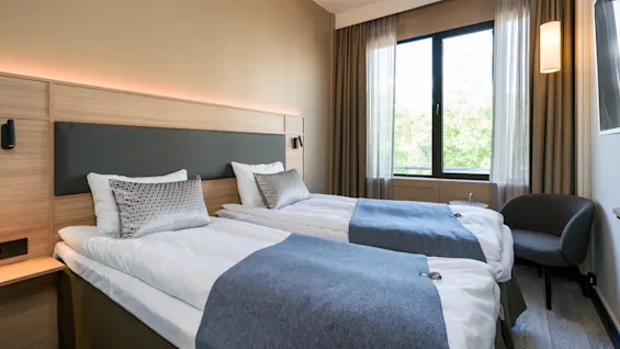 Two neatly made twin beds with gray throws are positioned side by side. A window with closed curtains illuminates the room, which features neutral walls and a modern chair.