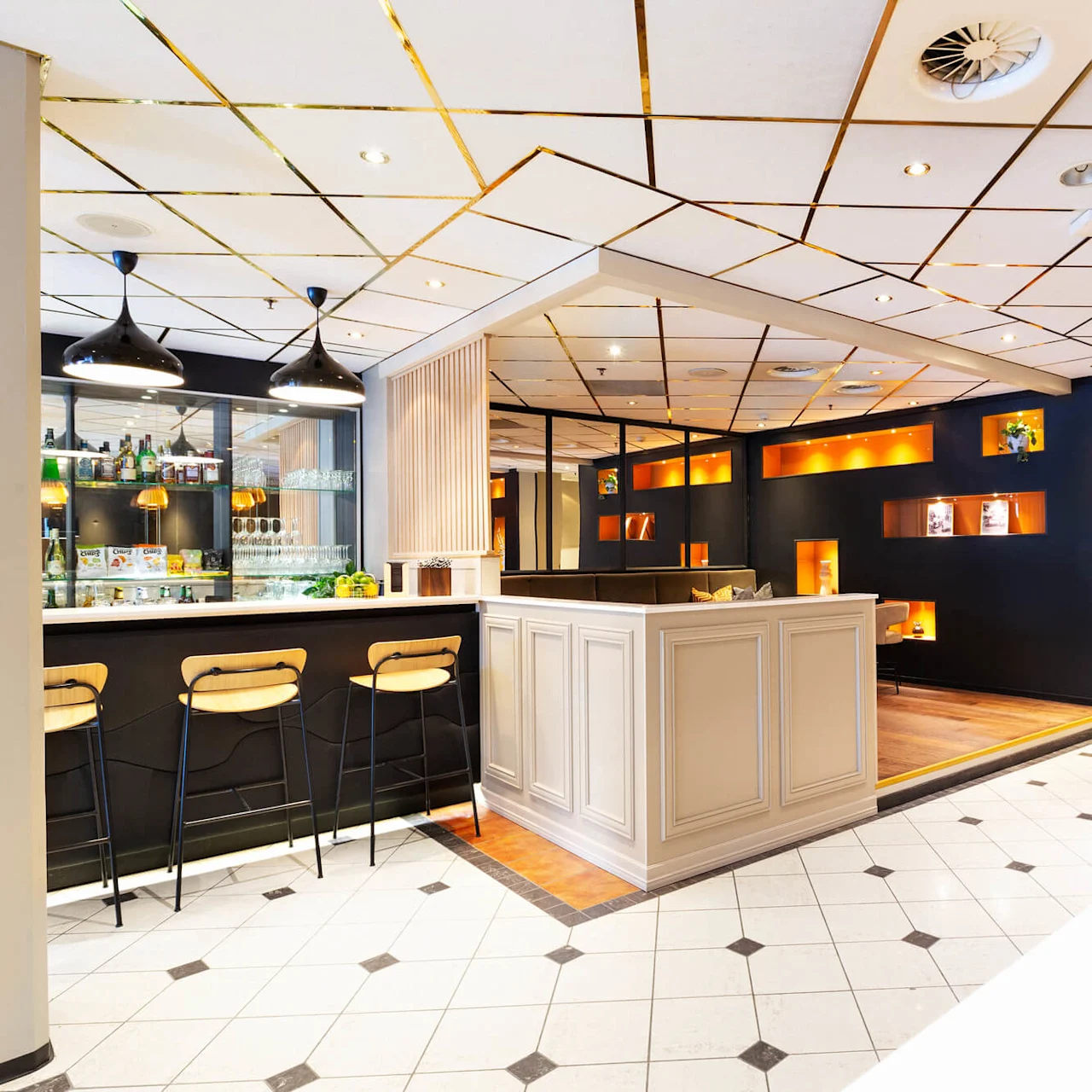 En moderne bar med stole står ved skranken under pendellamper, ved siden af et loungeområde med oplyste hylder. Omgivet af moderne designelementer og en poleret flisebelagt gulv.