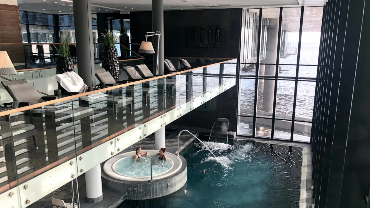 Et indendørs spa med en pool, hvor to personer slapper af, med udsigt til havet gennem store vinduer.