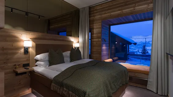 A cozy bedroom features a bed with patterned pillows and a blanket, illuminated by wall lamps. Large windows reveal a snowy mountain view, enhancing the wooden interior's warmth.