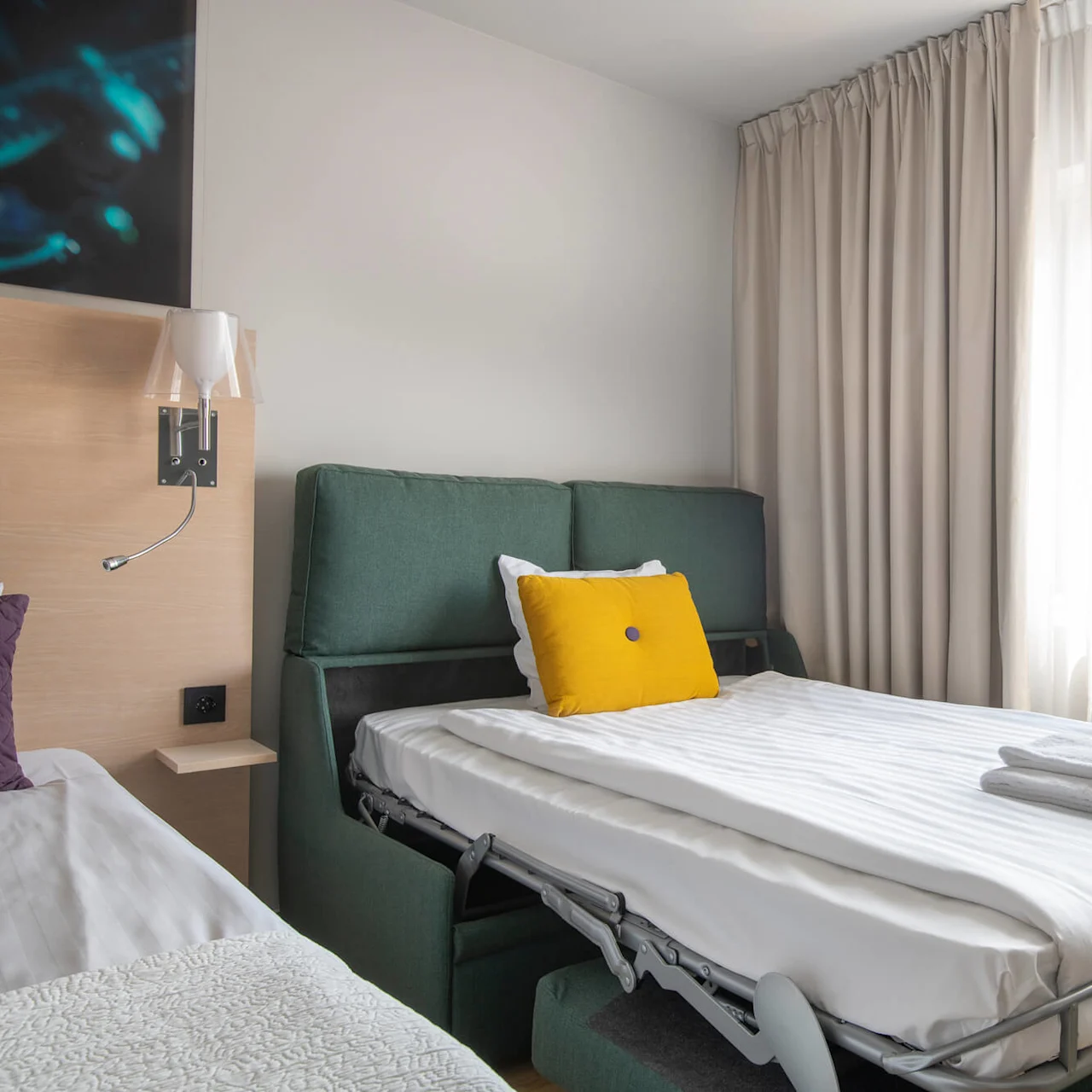 En säng med en gul kudde är utfälld från en grön soffa. Vita gardiner hänger bredvid ett fönster och ljusnar upp det moderna rummet med trädetaljer och en sänglampa.