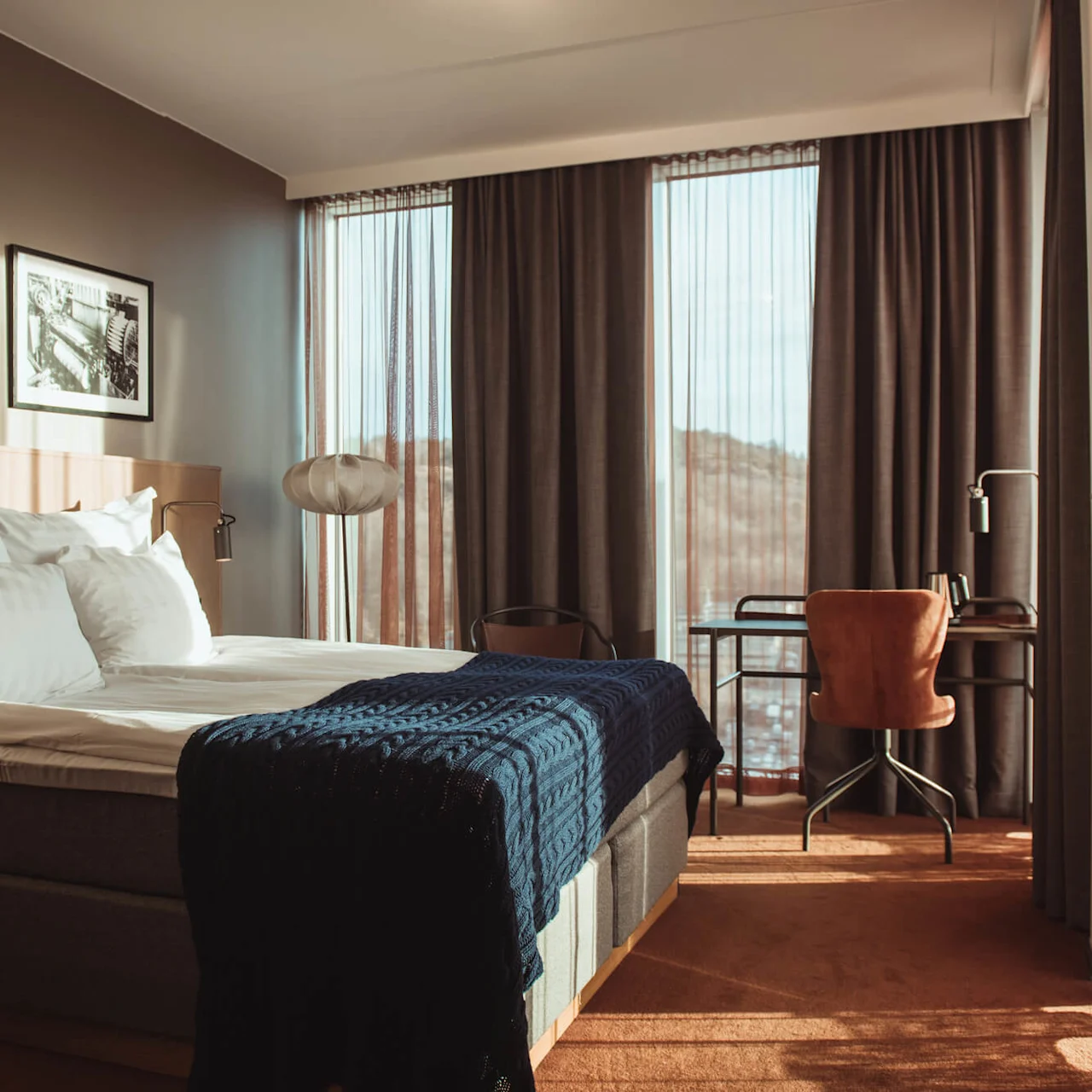 En noggrant bäddad säng med vita lakan och en blå filt står i ett hotellrum med stora fönster, en inramad bild på väggen och ett skrivbord med en stol.