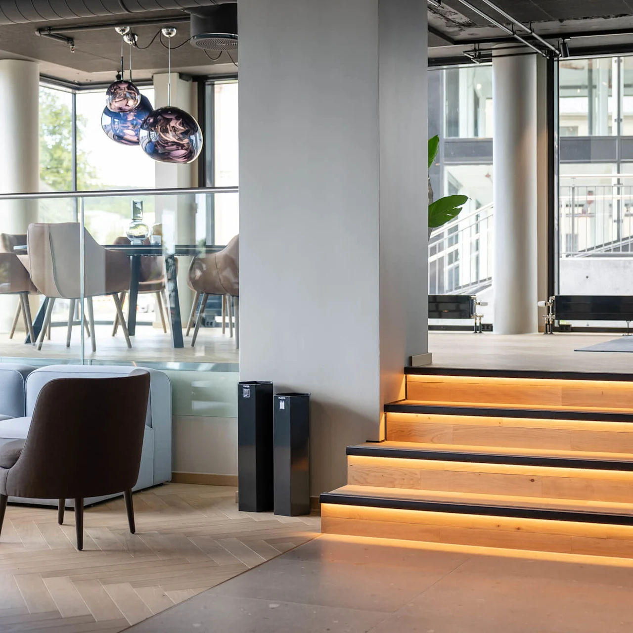 Moderne lounge med oplyste trappetrin, der fører op til et siddeområde med stole og borde, store vinduer og dekorative hængende lys, som skaber en rummelig og stilfuld atmosfære.