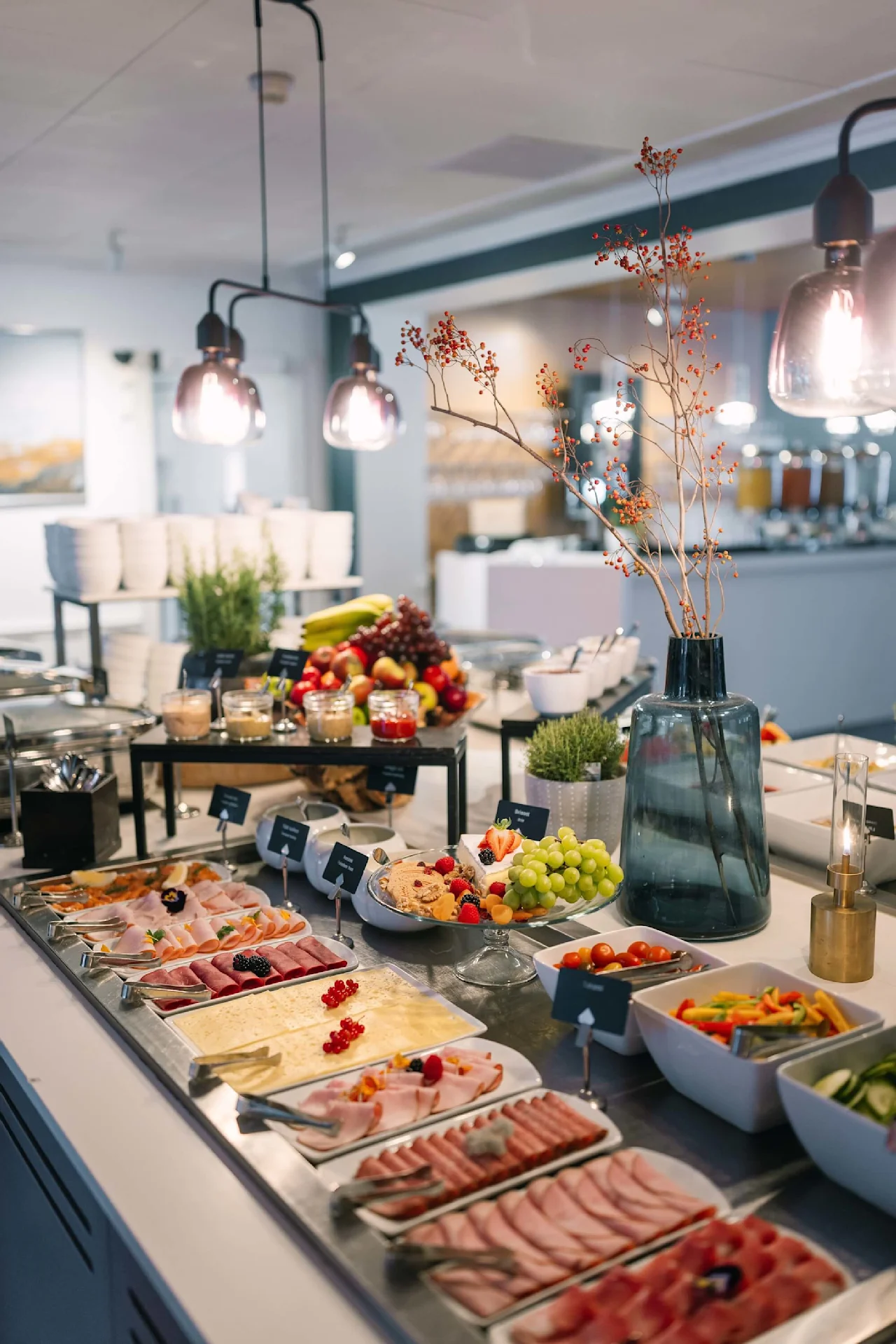 Buffetbord med forskellige pålæg, oste, frugt i et godt oplyst spiseområde under hængende lamper; ledsaget af dekorative grene i en vase. Etiketter inkluderer: "Skinka", "Rullader", "Rostbiff", "Salami".