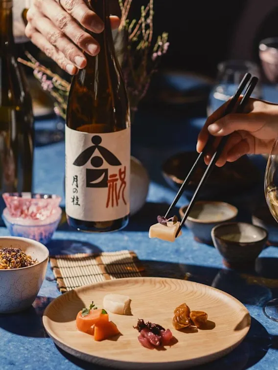 En hand använder ätpinnar för att ta sushi från en träplatta. En annan hand håller en sakeflaska med japansk text. Olika rätter och glas omger det blå bordet.