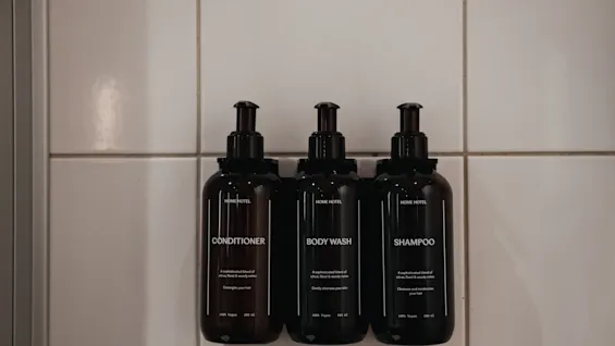 Tre mörka flaskor med balsam, duschtvål och schampo är monterade på en kaklad vägg, redo att användas.