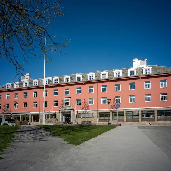 En stor, rødorange hotellbygning med mange vinduer under en klarblå himmel, med en asfaltert gangvei og grønt gress foran.