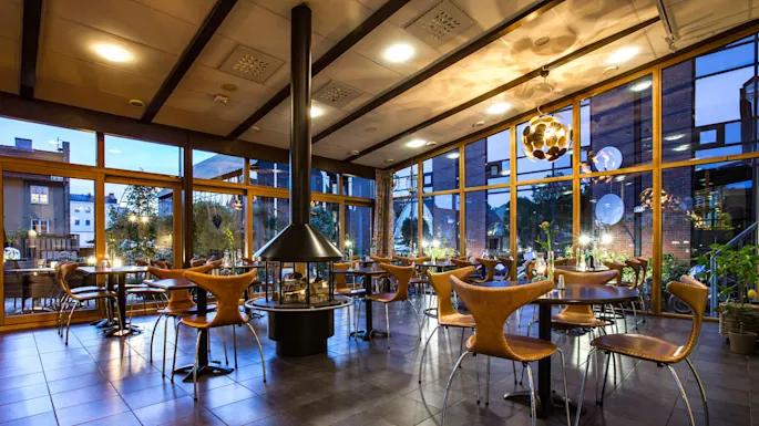 Et tomt, koselig kaféinteriør i skumring med stoler rundt små runde bord og peis i midten, vinduer fra gulv til tak viser kveldstimen utenfor.