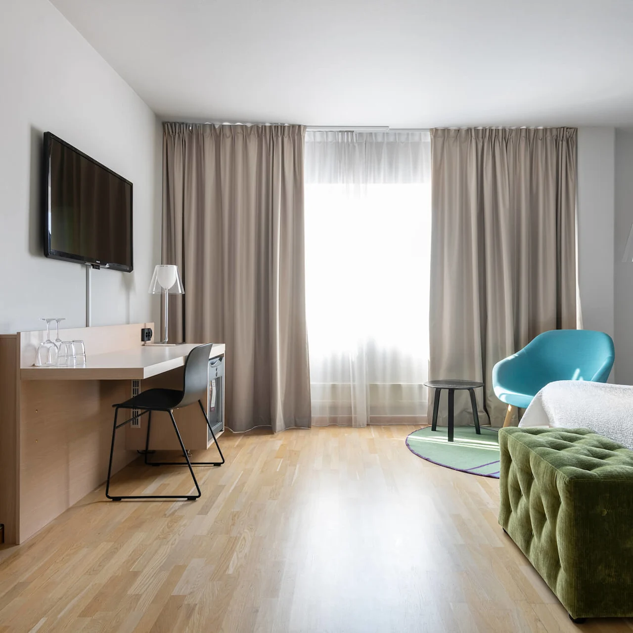 Ett prydligt organiserat hotellrum har en väggmonterad TV ovanför ett skrivbord med en stol, en säng med en grön ottoman och en blå stol nära gardinerade fönster.