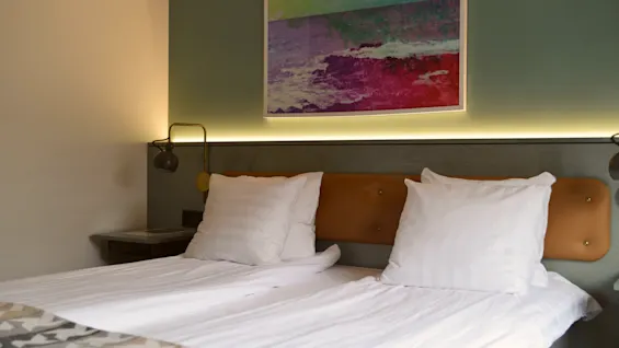 En pænt redt dobbeltseng med to hvide puder og et farverigt abstrakt maleri ovenfor, belyst af blød vægbelysning, i et moderne hotelværelse.