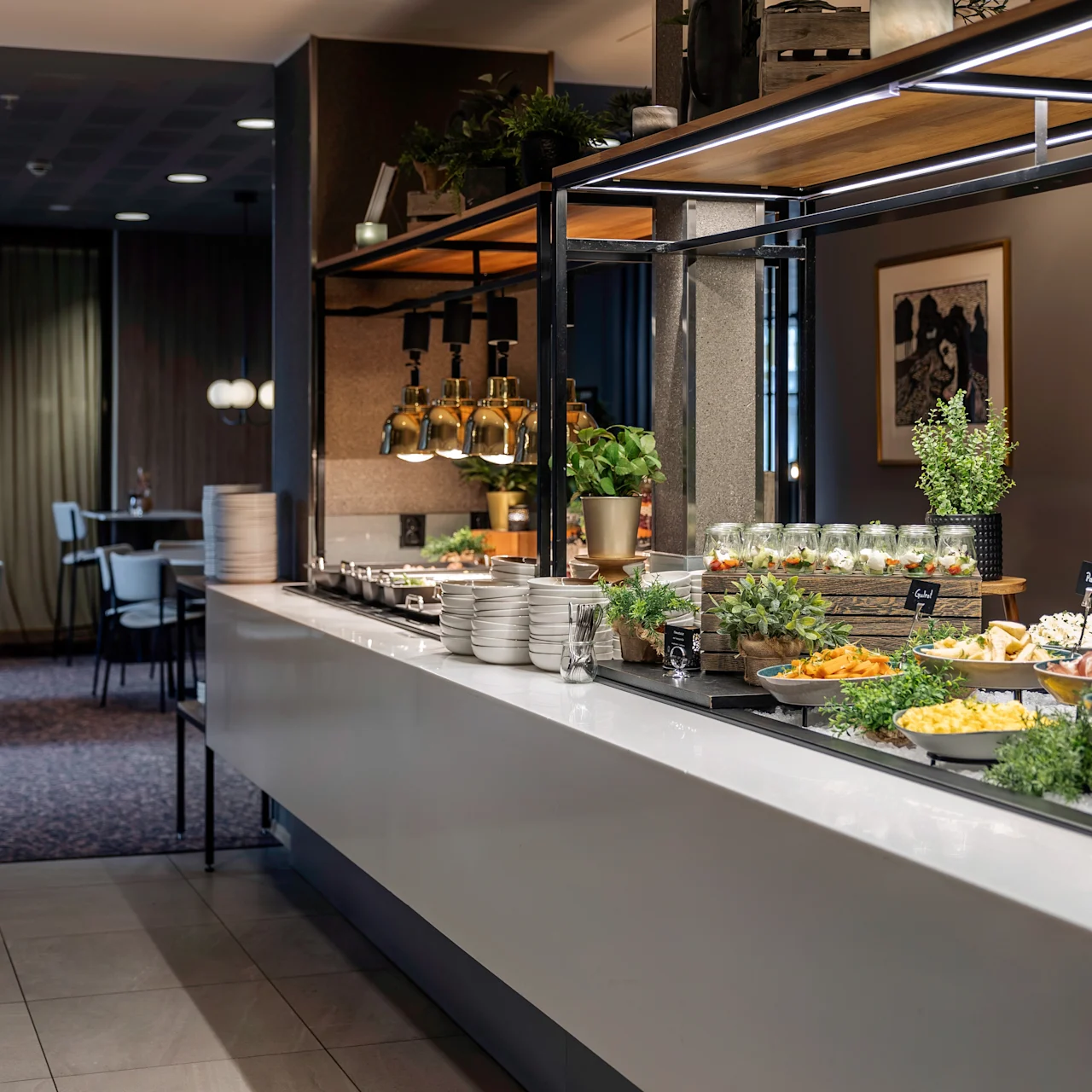 A buffet table displays various dishes, including salads in jars, roasted meats, and pasta. Plates and greenery decorate the counter in a modern dining area with tables and chairs.