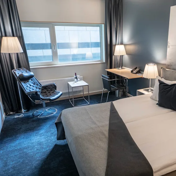 Säng prydligt bäddad med grått och vitt sänglinne; belägen i ett modernt hotellrum med en svart stol, litet bord med vin, skrivbord och mjuk belysning. Stort fönster ger gott om naturligt ljus.