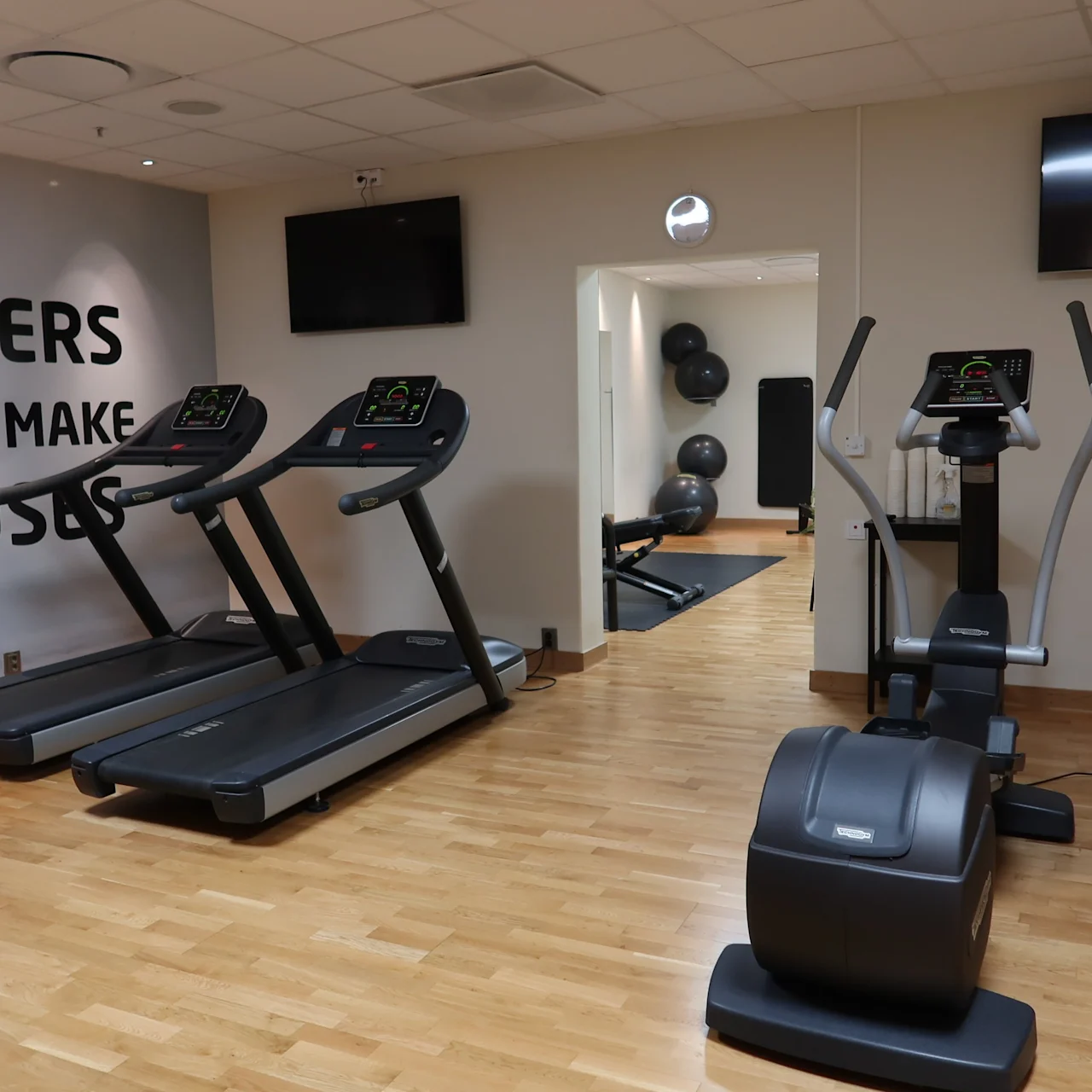 Treadmills and exercise bikes stand idle in a well-lit gym with wooden flooring. Walls display motivational text: "WINNERS DO NOT MAKE EXCUSES". Balls are visible through an interior doorway.