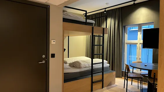 Moderni hotellihuone, jossa on kerrossänky, ruokapöytä tuoleineen ja ikkuna tummilla verhoilla.