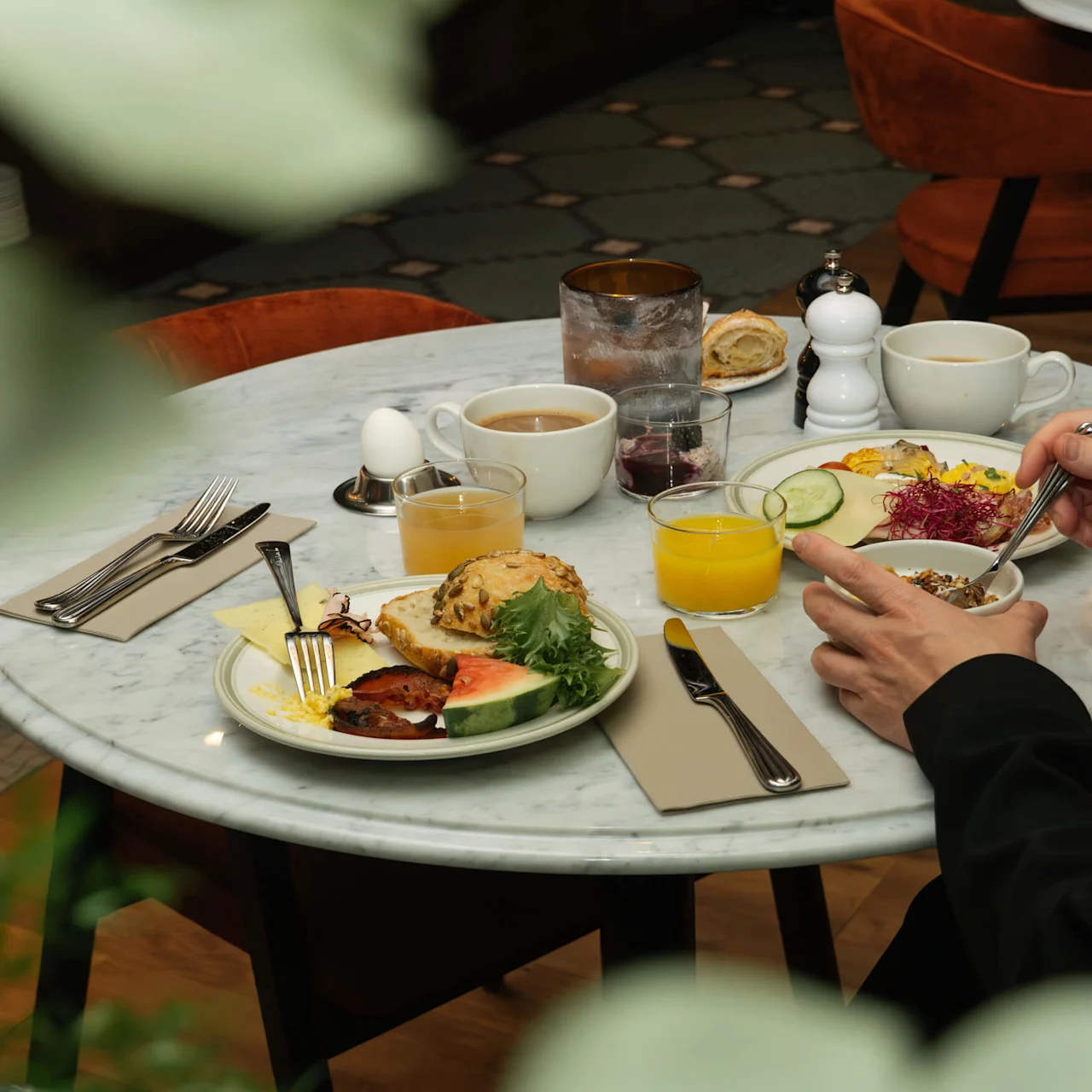 Breakfast plates with bread, fruit, and eggs are on a marble table. Someone is eating with utensils. Coffee cups, juice, and cutlery are also present, surrounded by orange chairs.