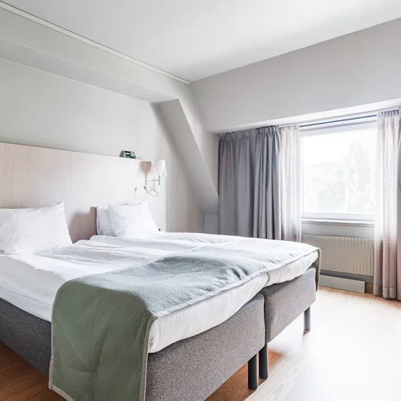 Ljust hotellrum med två bäddade sängar bredvid varandra och ett stort fönster med grå gardiner.