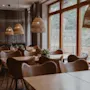 A cozy restaurant dining area with wooden tables, leather chairs, and woven pendant lights. Large windows overlook an outdoor space.