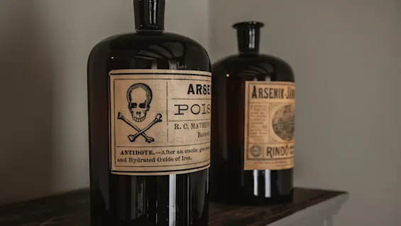 Two dark glass bottles with vintage labels, one featuring a skull and crossbones. Labels read ARSE POIS, ANTIDOTE, ARSENIK-J.