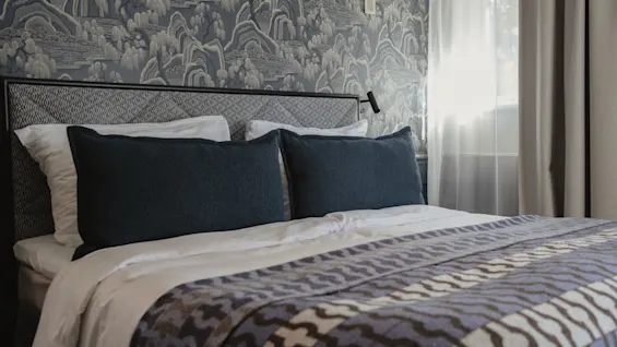 Ett bekvämt sovrum med en prydligt bäddad säng, mörka kuddar och en mönstrad filt. Solljus silas in genom fönstret.