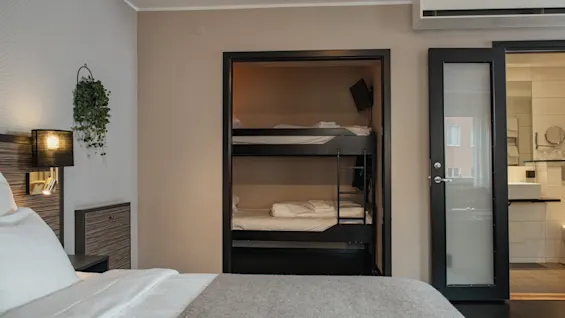 Hotellihuone, jossa on mukava sänky, sisäänrakennettu kerrossänky lisävieraille ja valoisa kylpyhuone avoimen oven kautta.